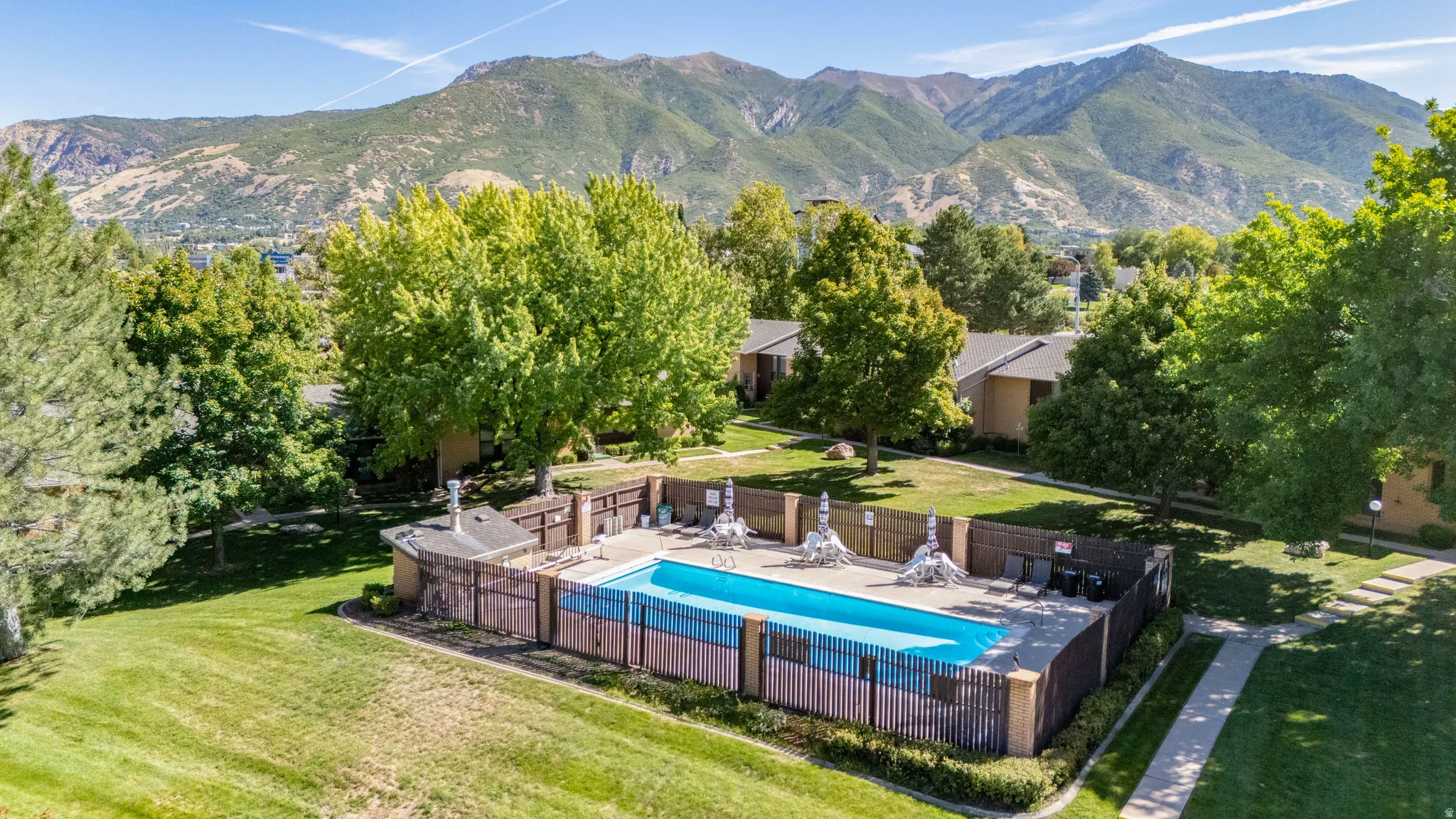 Community swimming pool with surrounding fencing,  lawn area, and mature trees, set against a mountainous backdrop