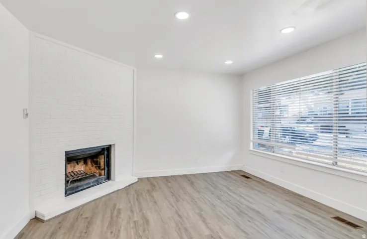 White painted brick fireplace, wood-finish flooring, recessed lighting, expansive window with blinds, and white trim