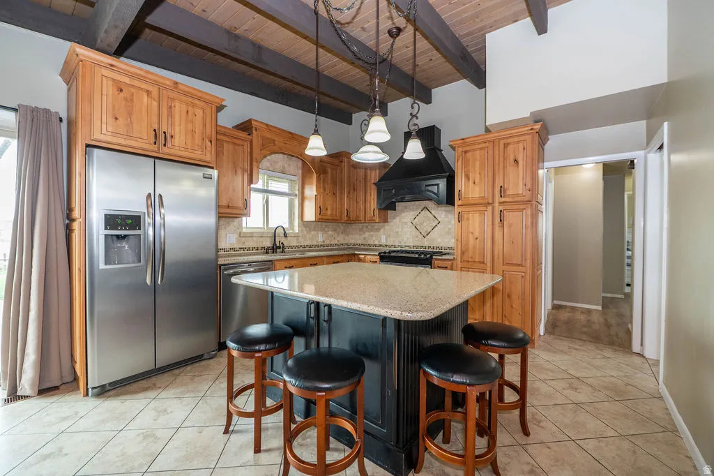 Vaulted wood-plank ceiling with exposed beams, stainless steel appliances, wood cabinetry, tile flooring, and a central island with a solid surface countertop