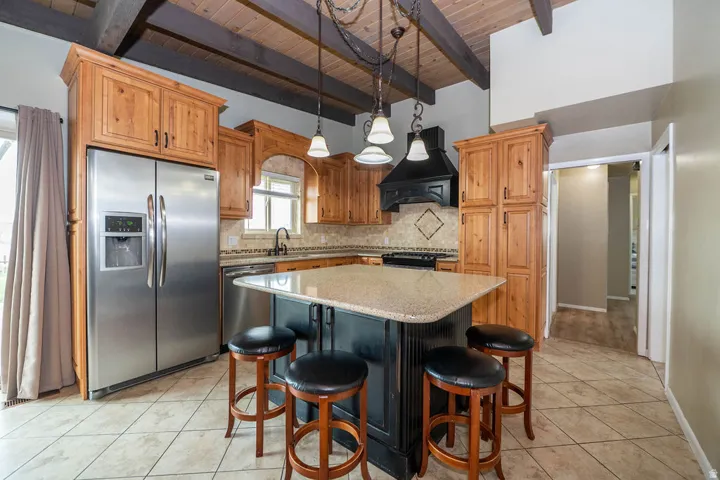Vaulted wood-plank ceiling with exposed beams, stainless steel appliances, wood cabinetry, tile flooring, and a central island with a solid surface countertop