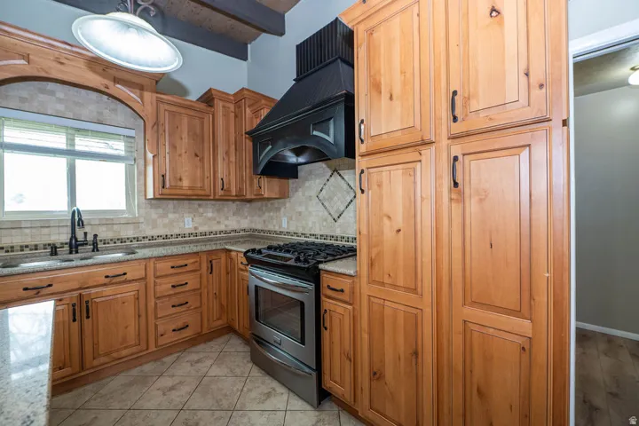 Wood cabinetry with black hardware, stainless steel range with black hood, tile flooring, stone countertops, and tile backsplash