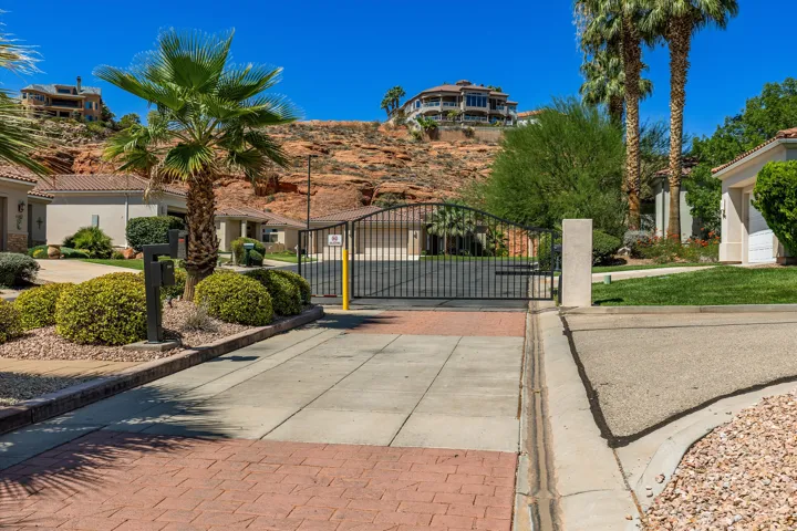 Gated community entrance featuring a paved driveway, manicured landscaping, and mature palm trees