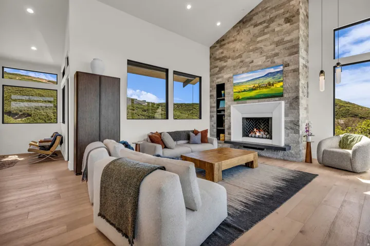 Vaulted ceilings with recessed lighting, stone-clad fireplace with white surround, light wood-finish flooring, and large windows with black frames