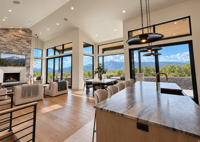 Spacious open-concept living area featuring high vaulted ceilings, large pane windows, wood-finish flooring, a stone-clad fireplace, and an integrated kitchen island with a basin sink