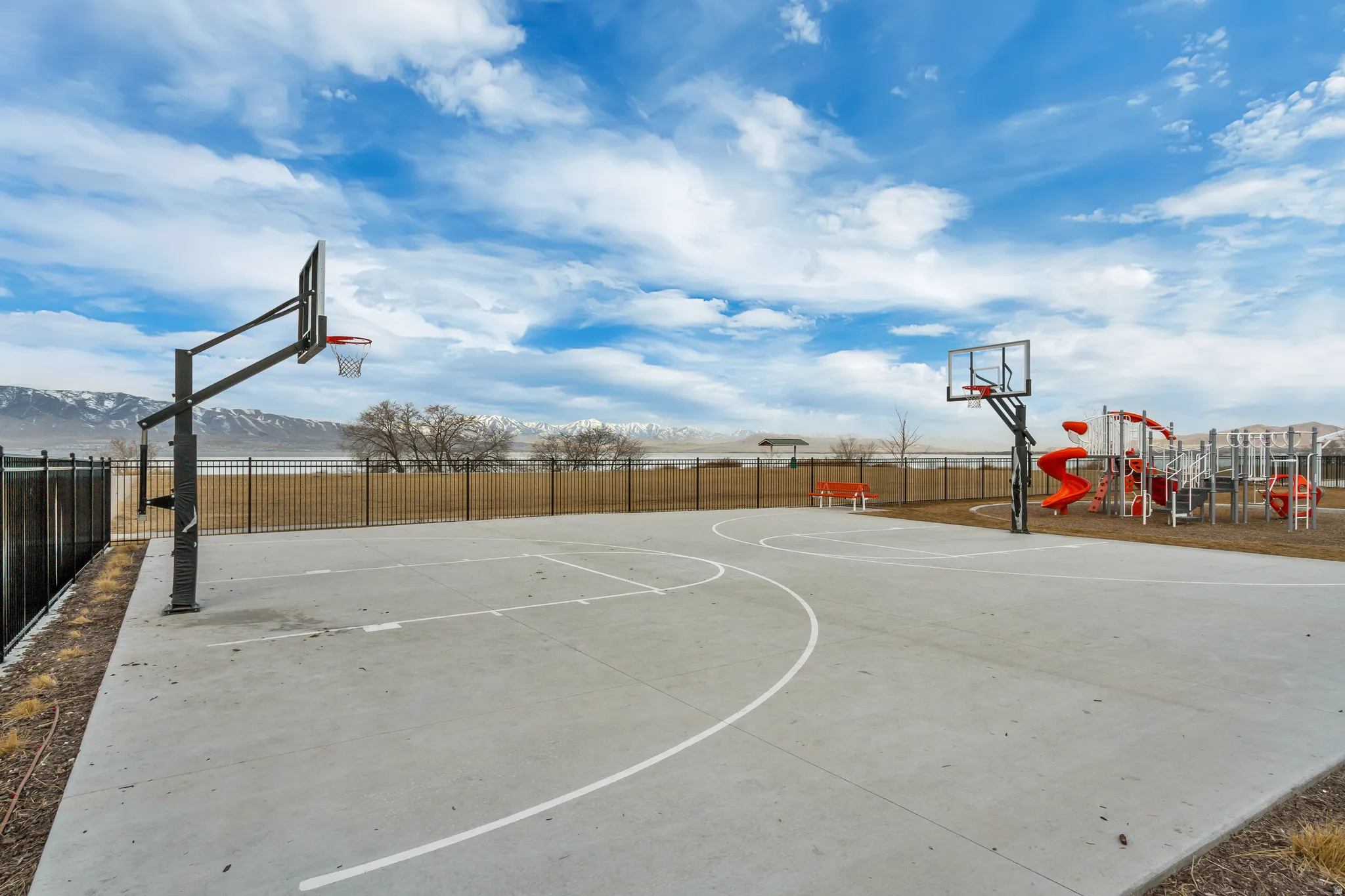 Outdoor concrete basketball court with two hoops, adjacent playground with slides and climbing structures, and a perimeter fence