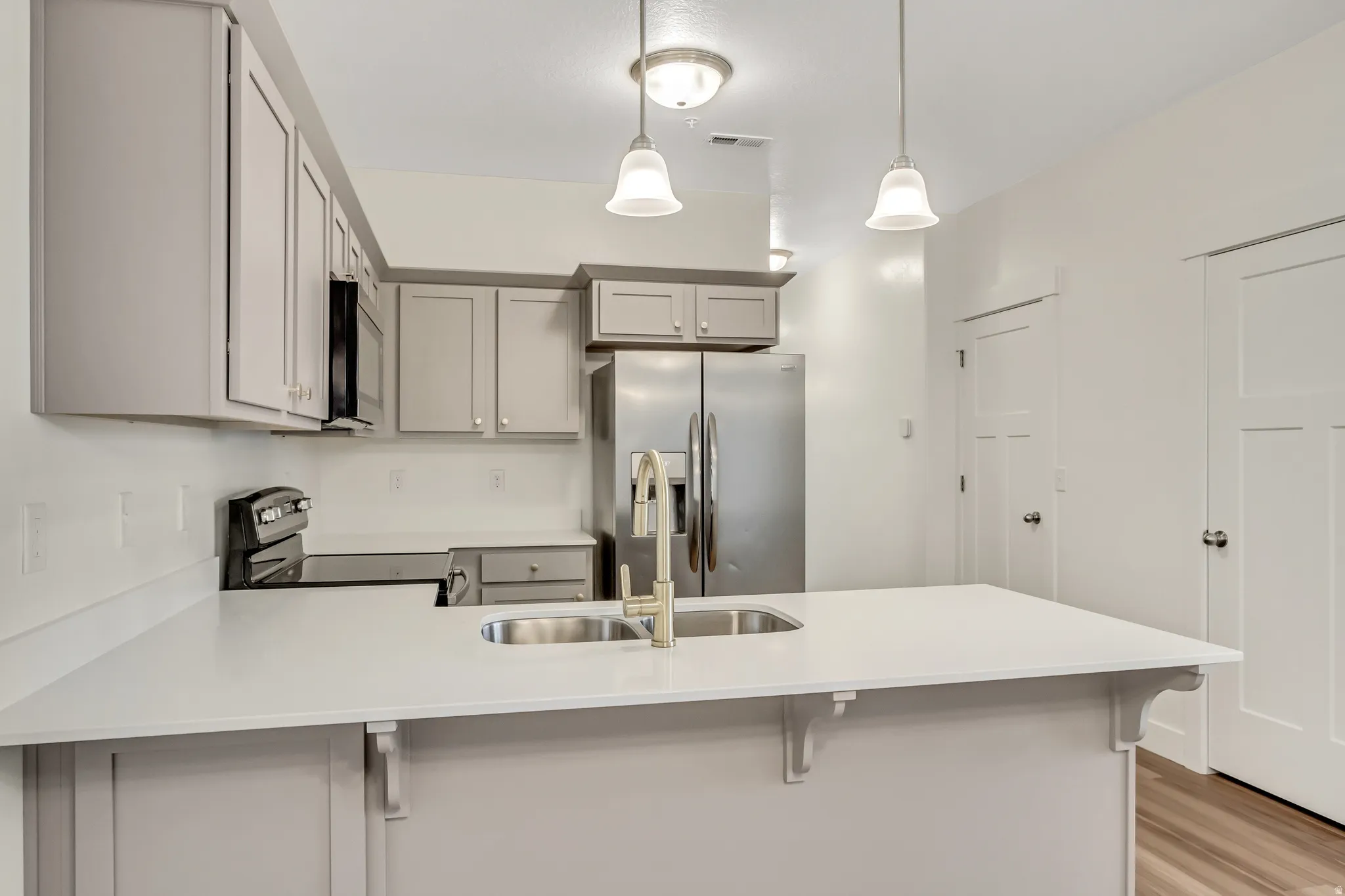 Kitchen featuring light gray cabinetry, stainless steel appliances, a center island with a double basin sink, and pendant lighting