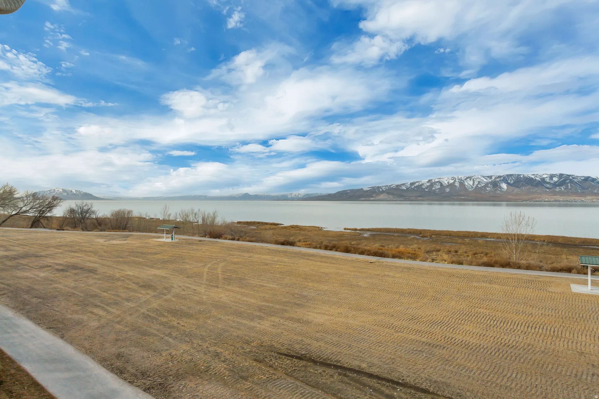Expansive open land with a paved pathway, bordered by a body of water and distant snow-capped mountains