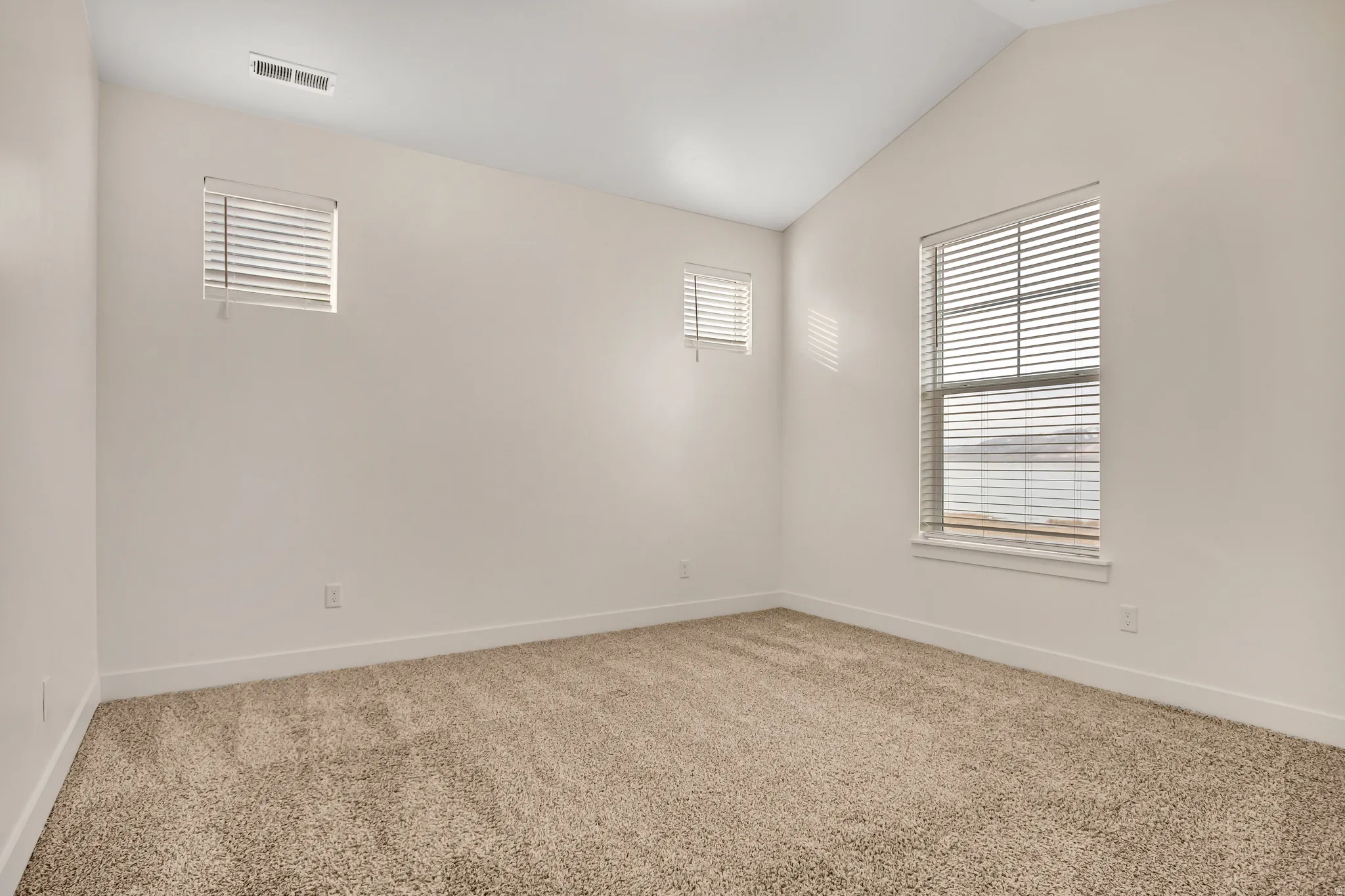 Vaulted ceiling, two windows with blinds, light-colored carpet, and white walls
