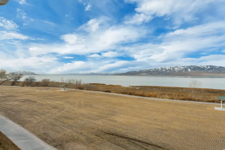 Expansive open land with a paved pathway, bordered by a body of water and distant snow-capped mountains