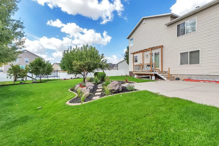 Expansive green lawn with a rock garden and stepping stones, featuring mature trees and a white privacy fence, complemented by a raised wood deck with a pergola and steps down to a concrete patio