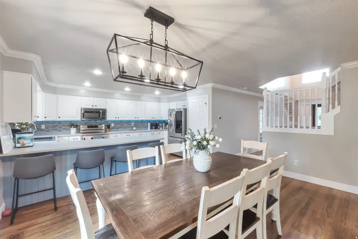 Open-concept dining area featuring wood-finish flooring, a linear chandelier, and access to a kitchen with white cabinetry and a tile backsplash