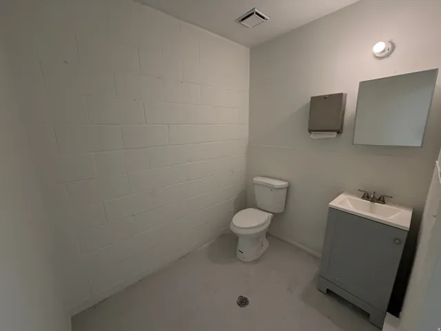 White painted cinder block walls, white toilet, single vanity with sink, wall-mounted mirror, and ceiling-mounted exhaust fan