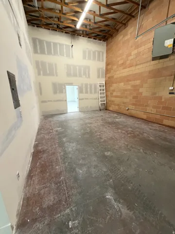 Exposed wood beam ceiling with fluorescent lighting, brick accent wall, concrete flooring, and a drywall finish