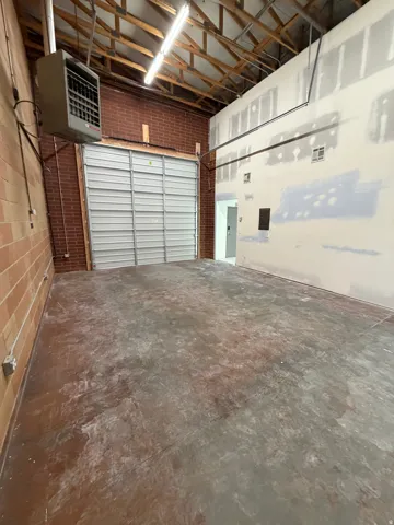 Exposed wood beam ceiling with fluorescent lighting, brick accent wall, mounted utility heater, and concrete flooring