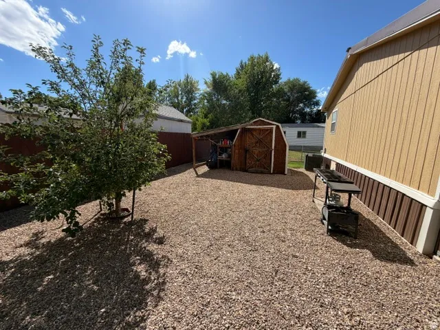 Gravel yard featuring a young tree, a brown privacy fence, a wooden shed with an open lean-to, and light-colored siding