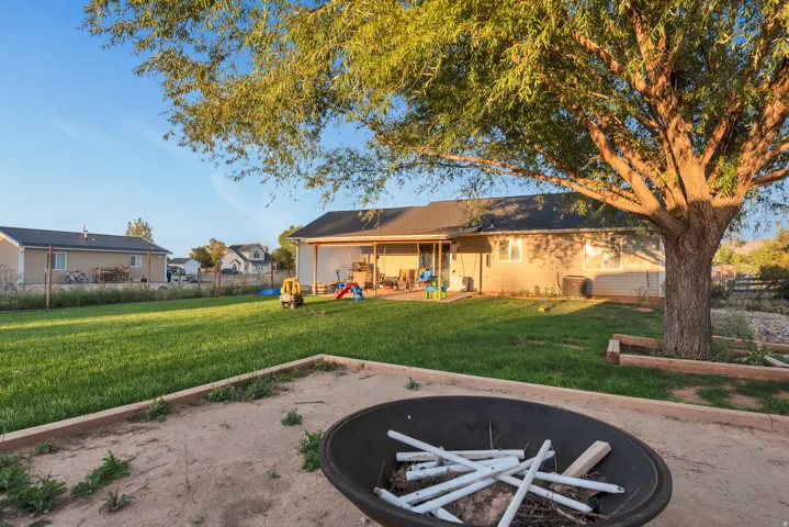 Expansive backyard featuring a manicured lawn, established shade tree, covered patio, and fire pit