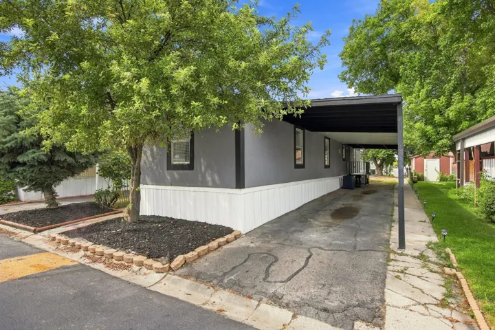 Grey siding with white trim, covered carport, landscaping with mature trees, and a paved driveway