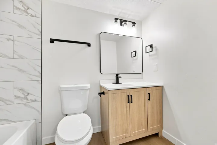 White subway-style tile shower surround, contemporary vanity with a single basin sink, matte black fixtures, framed mirror, and wood-finish flooring
