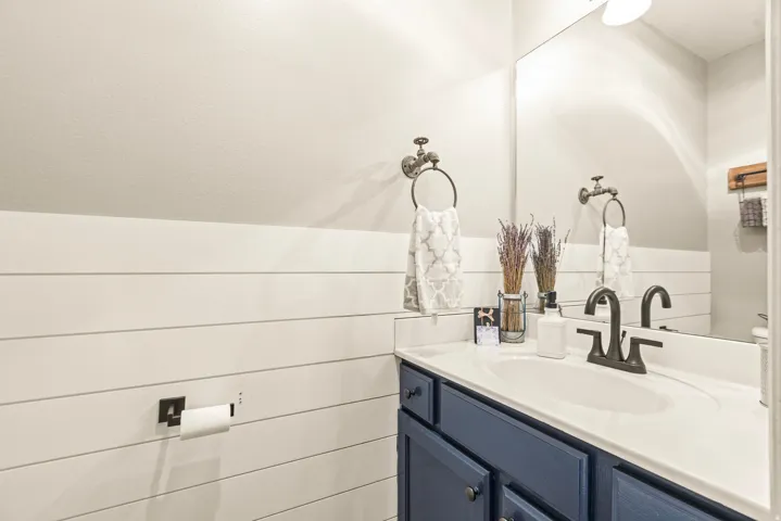 Shiplap accent wall, white countertop, dark blue vanity with cabinet storage, integrated sink, and dark bronze faucet