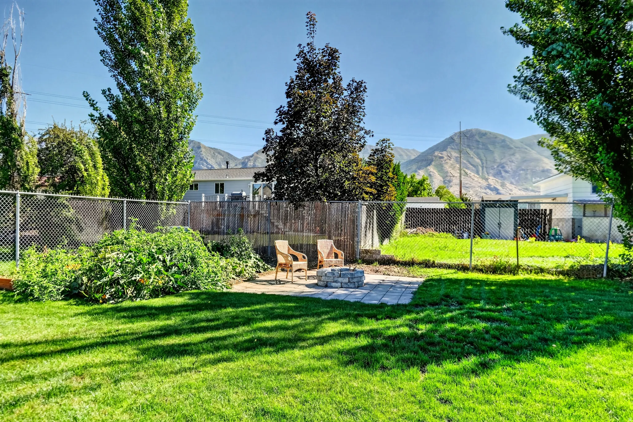 Expansive backyard featuring a manicured lawn, stone paver patio, stone fire pit, and chain link fencing