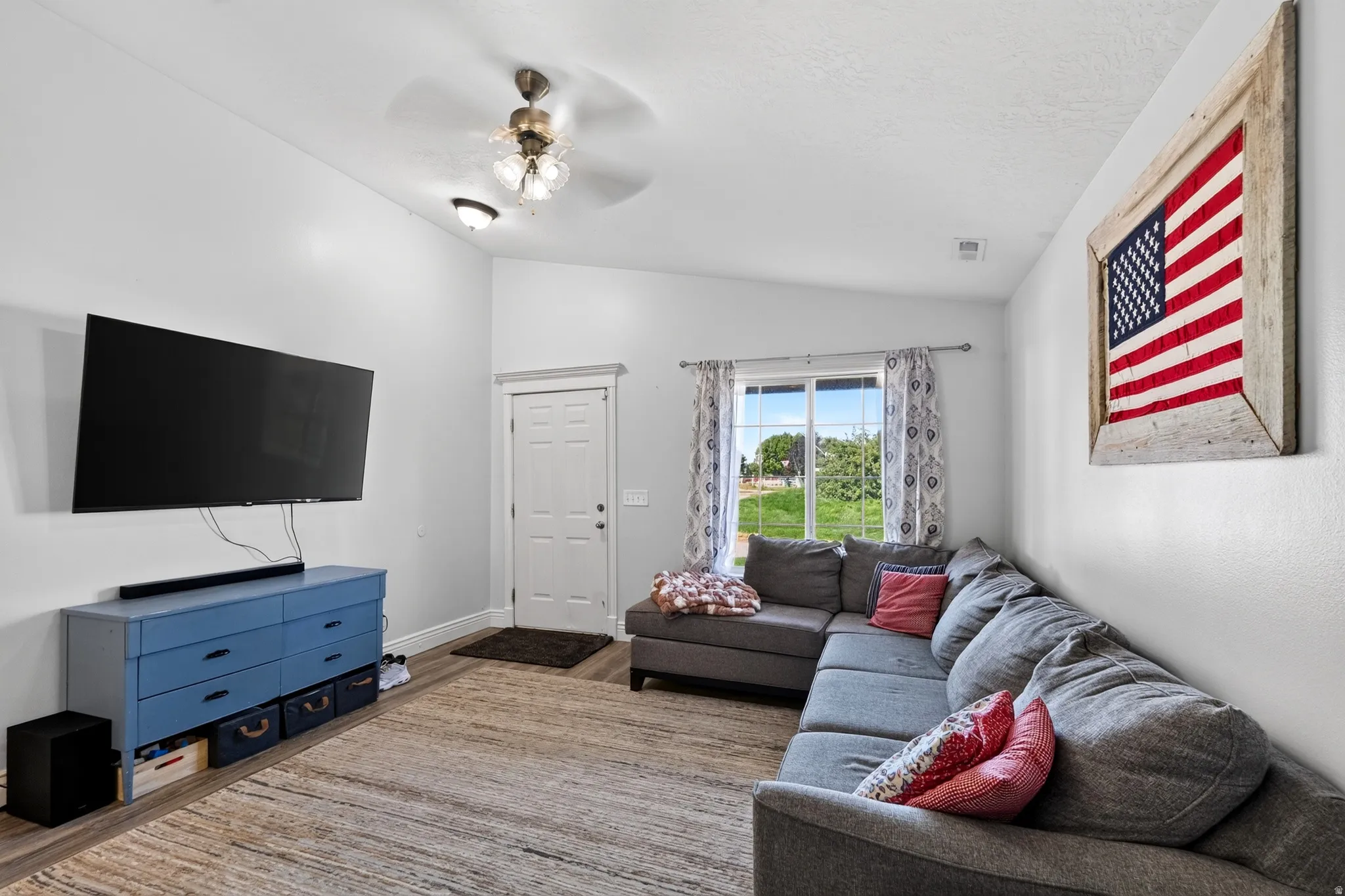 Vaulted ceiling with ceiling fan and integrated lighting, wood-finish flooring, white paneled door, and a large window