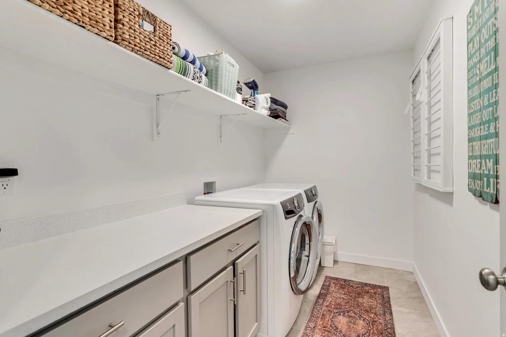 Laundry area featuring an expansive countertop, built-in cabinetry with silver hardware, wall-mounted shelving, and a window with white shutters