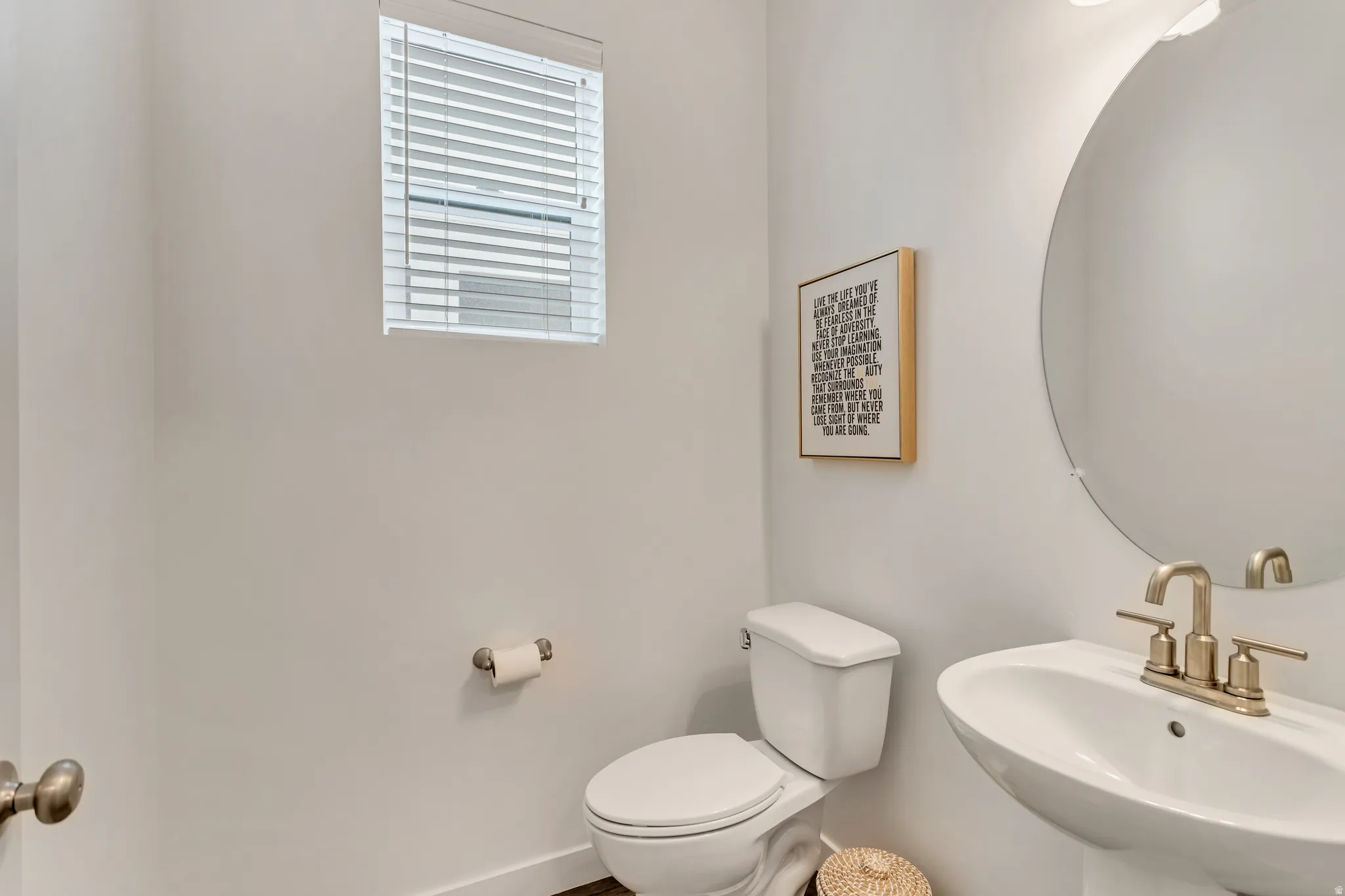 Pedestal sink with brass-finish faucet, circular mirror, and window with blinds