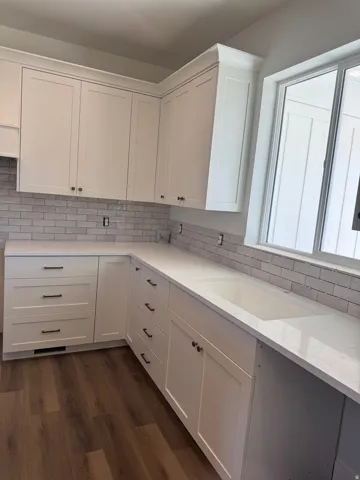 White shaker-style cabinetry with gold-tone hardware, subway tile backsplash, solid surface countertops, integrated sink, and wood-finish flooring