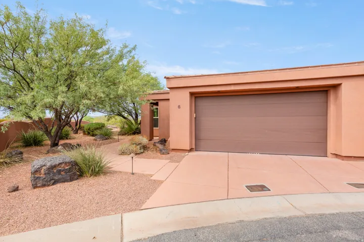 Stucco exterior with a two-car garage, concrete driveway, and xeriscape landscaping featuring mature trees and decorative rocks