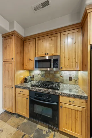 Light wood cabinetry, stainless steel microwave, black gas range, built-in oven, and multi-toned stone tile flooring
