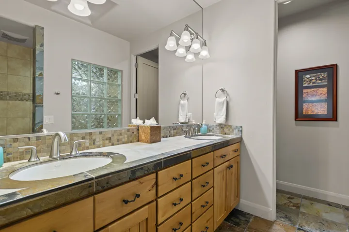 Double vanity with integrated sinks, stone countertops, natural wood cabinetry, mosaic tile backsplash, and overhead vanity lighting