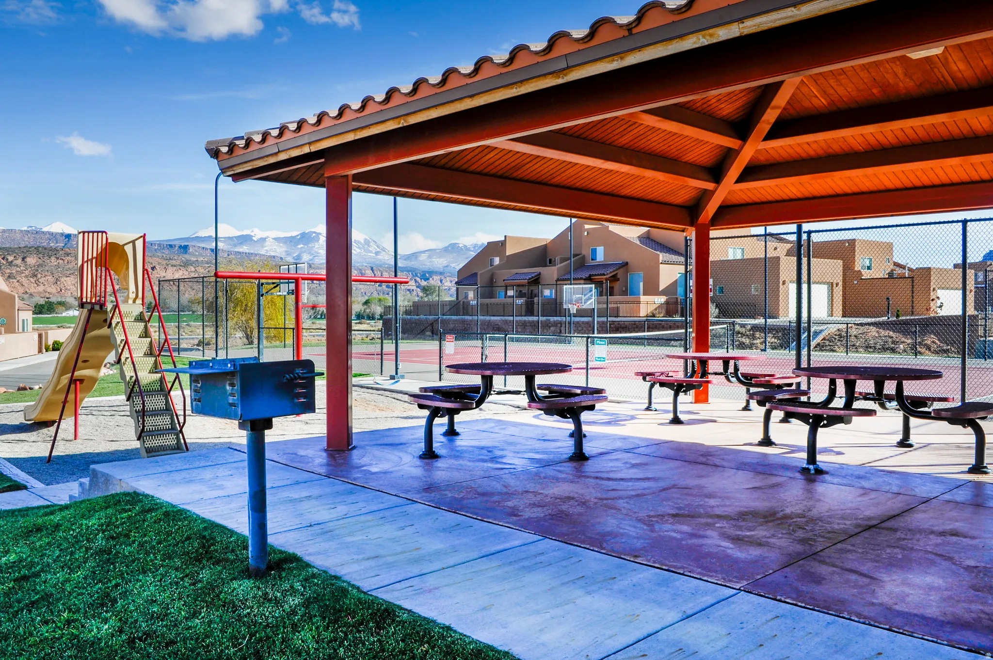 Shaded outdoor pavilion with picnic tables, adjacent to a playground and sports court, with desert mountain scenery