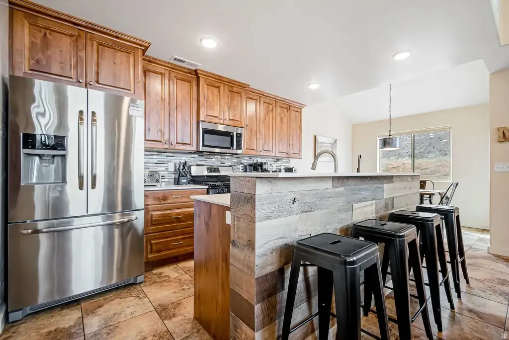 Stainless steel appliances, wood cabinetry, tile flooring, kitchen island with wood paneling, and pendant lighting