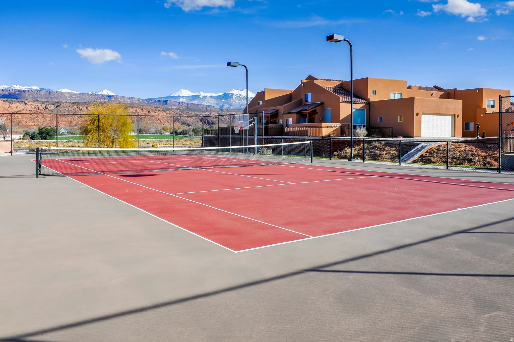 Red surface tennis court with perimeter fencing, street lighting, and a stucco-exterior building