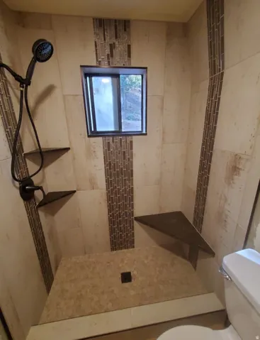Tiled shower with a handheld showerhead, built-in corner shelves, and a window