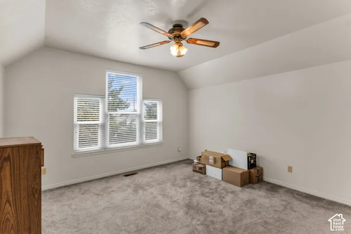 Carpeted room with a vaulted ceiling, ceiling fan with light fixture, and a multi-panel window unit featuring blinds