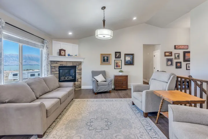 Vaulted ceiling, stone-surround fireplace with mantel, wood-finish flooring, recessed lighting, and a prominent picture window