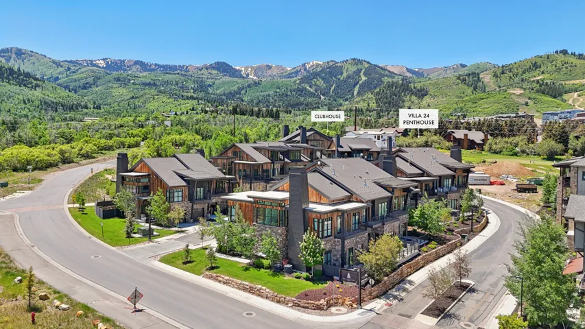 Mountain architecture featuring stone and wood siding, multiple private balconies, a winding paved road, and surrounding mature trees with a mountain backdrop