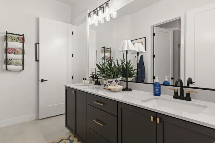 Dual undermount sinks with matte black and gold-tone fixtures, white countertops, dark cabinetry with gold-tone hardware, and an expansive mirror with strip lighting