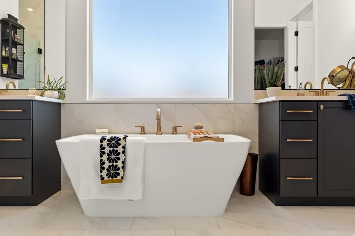 Freestanding soaking tub with floor-mounted faucet, dual vanities with dark cabinetry and gold-tone hardware, and large format tiled flooring