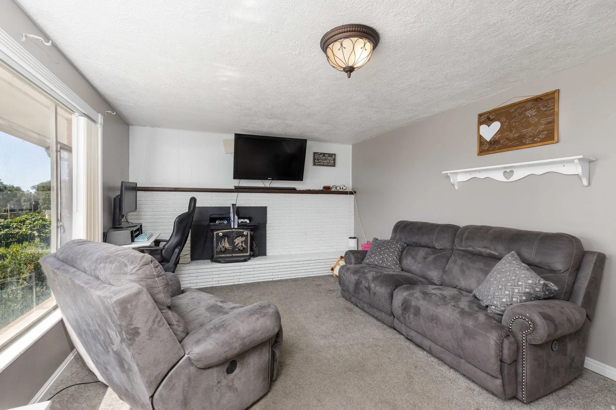 Spacious living area featuring a white brick fireplace with a dark wood mantel, large windows, and a ceiling-mounted light fixture