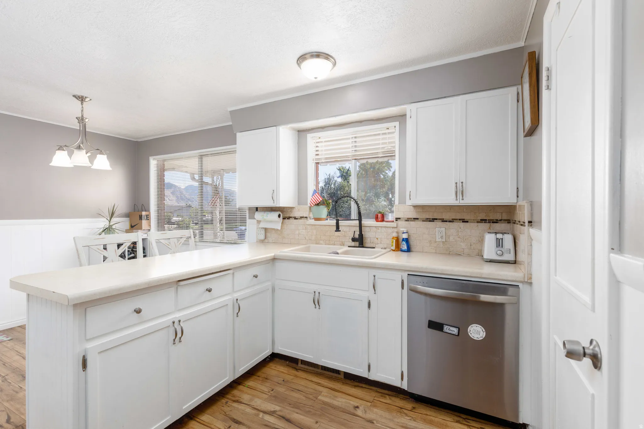 White cabinetry with brushed nickel hardware, L-shaped countertop, stainless steel dishwasher, wood-finish flooring, and a tile backsplash
