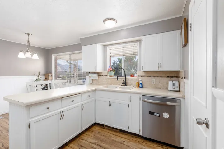 White cabinetry with brushed nickel hardware, L-shaped countertop, stainless steel dishwasher, wood-finish flooring, and a tile backsplash