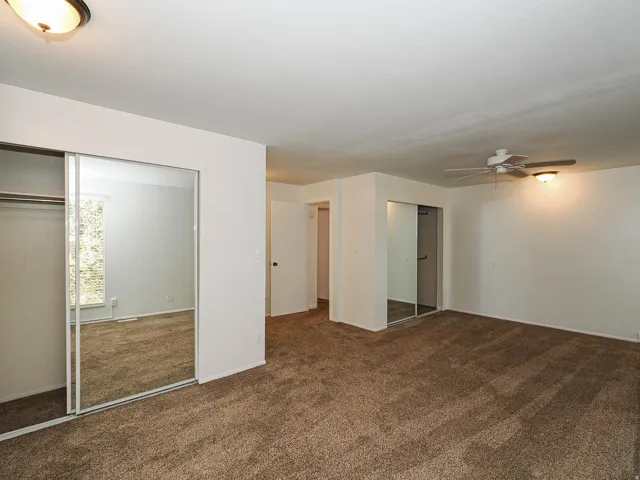 Spacious room featuring neutral carpeting, two sets of mirrored sliding closet doors, and a ceiling fan