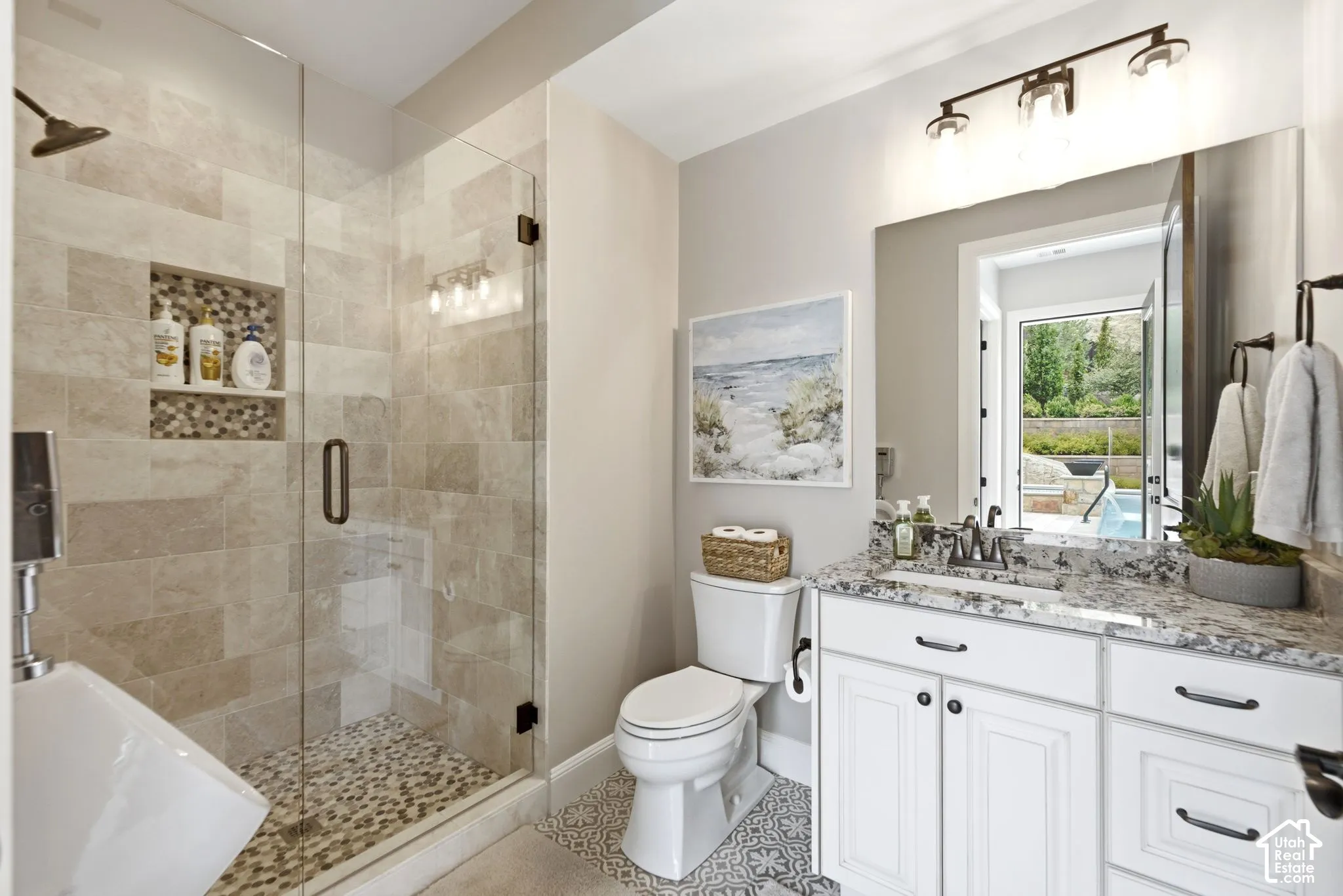Walk-in shower with natural stone tile surround and mosaic floor, glass enclosure, white vanity with stone countertop, and decorative floor tile