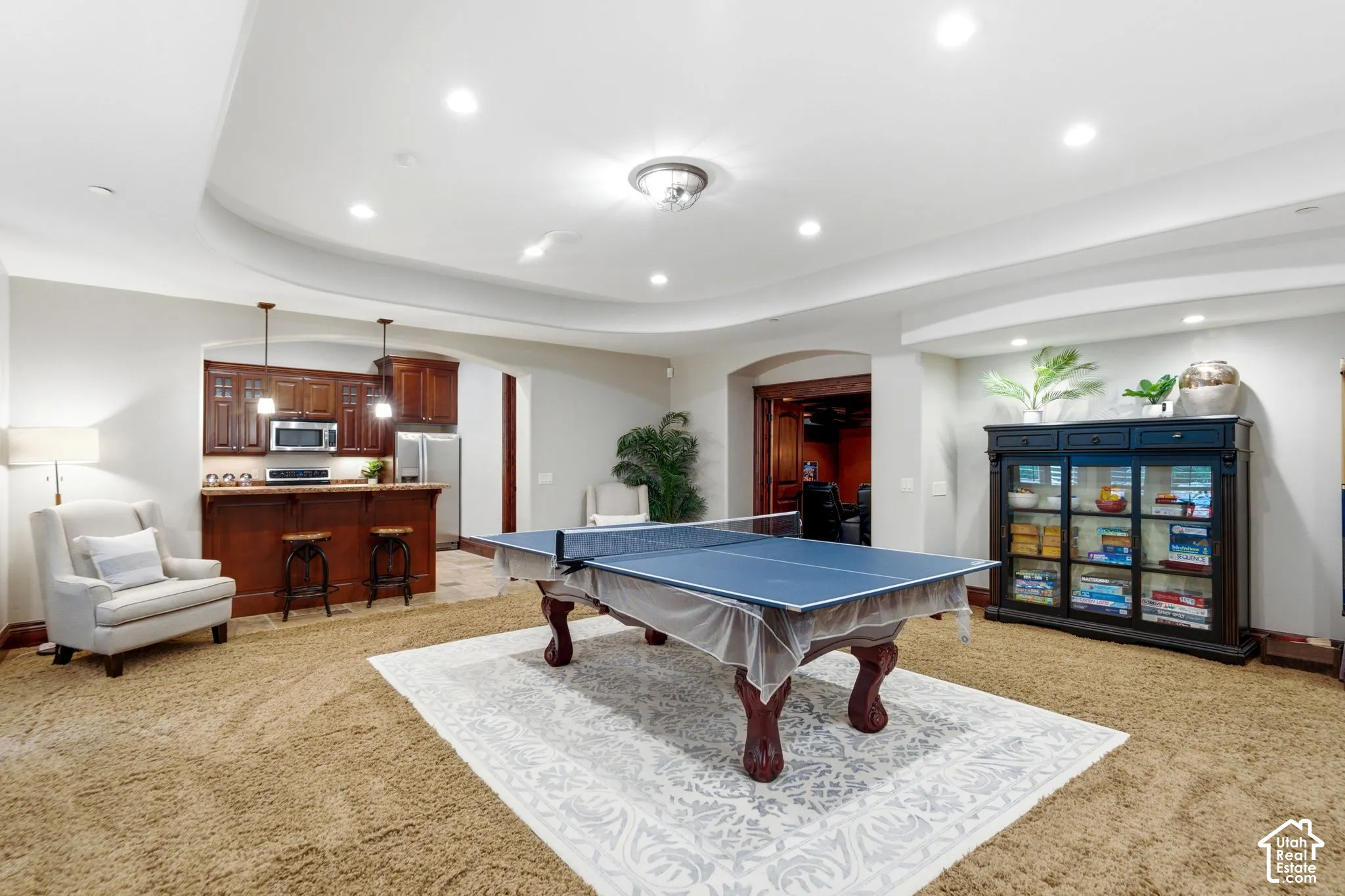 Recessed lighting illuminating a spacious carpeted area with an integrated wet bar featuring wood cabinetry and stainless steel appliances