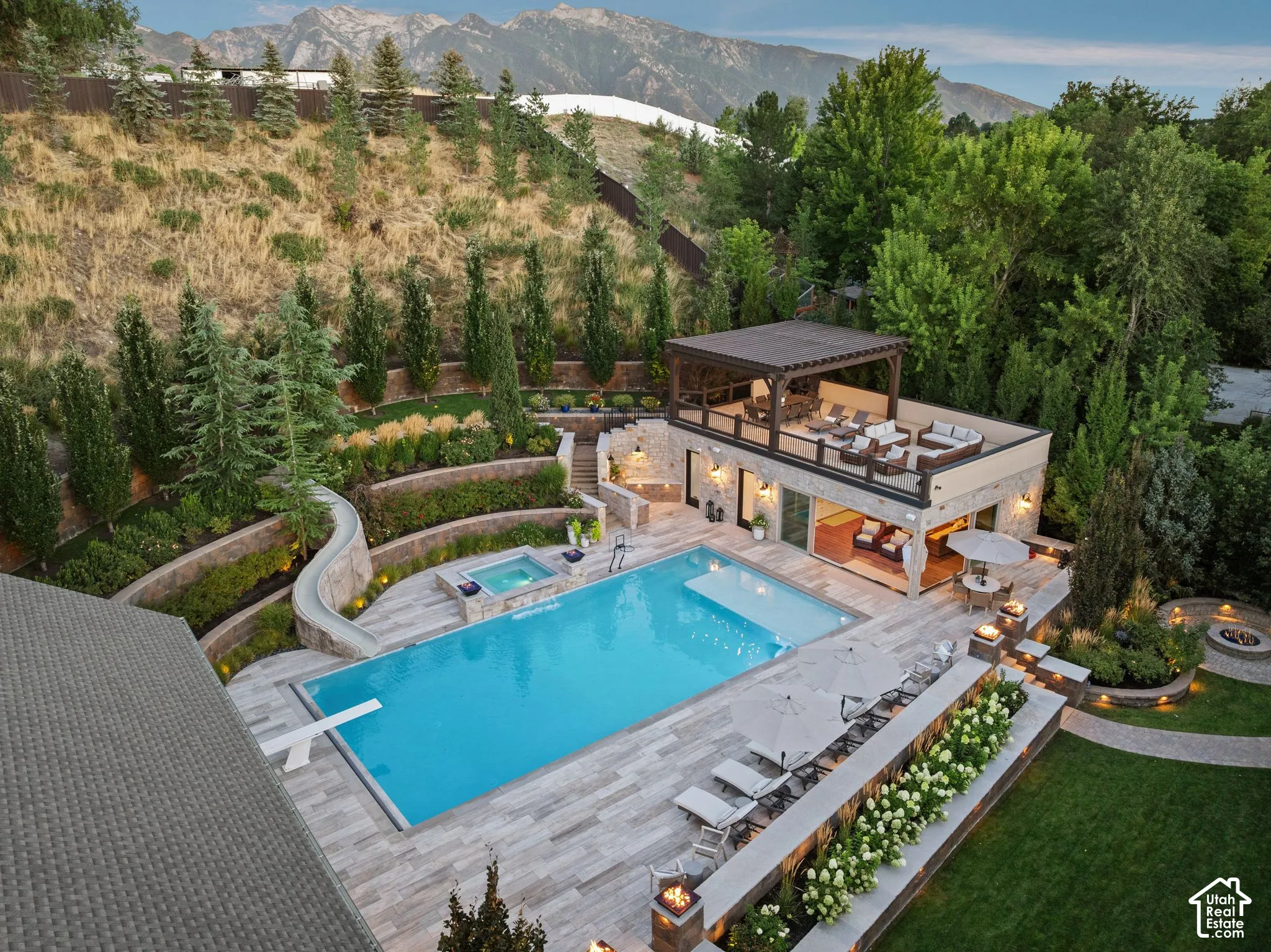 Terraced landscaping with mature trees, expansive swimming pool with diving board, integrated spa, dedicated outdoor structure with an upper-level pergola and lower-level covered patio, and multiple built-in fire features