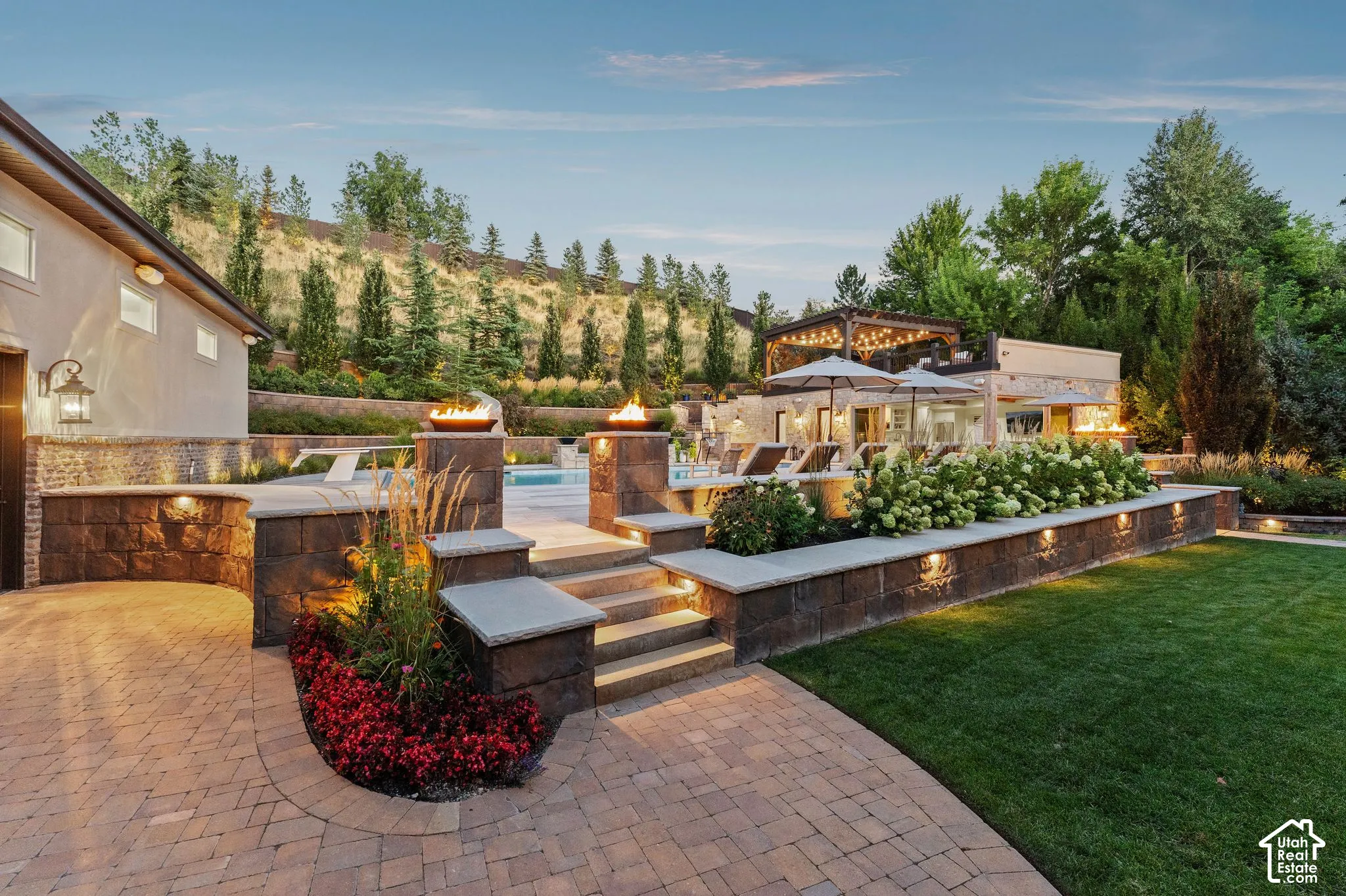 Paver driveway and walkways, stone retaining walls with integrated lighting, built-in planters, manicured lawn, and a swimming pool with adjacent pergola