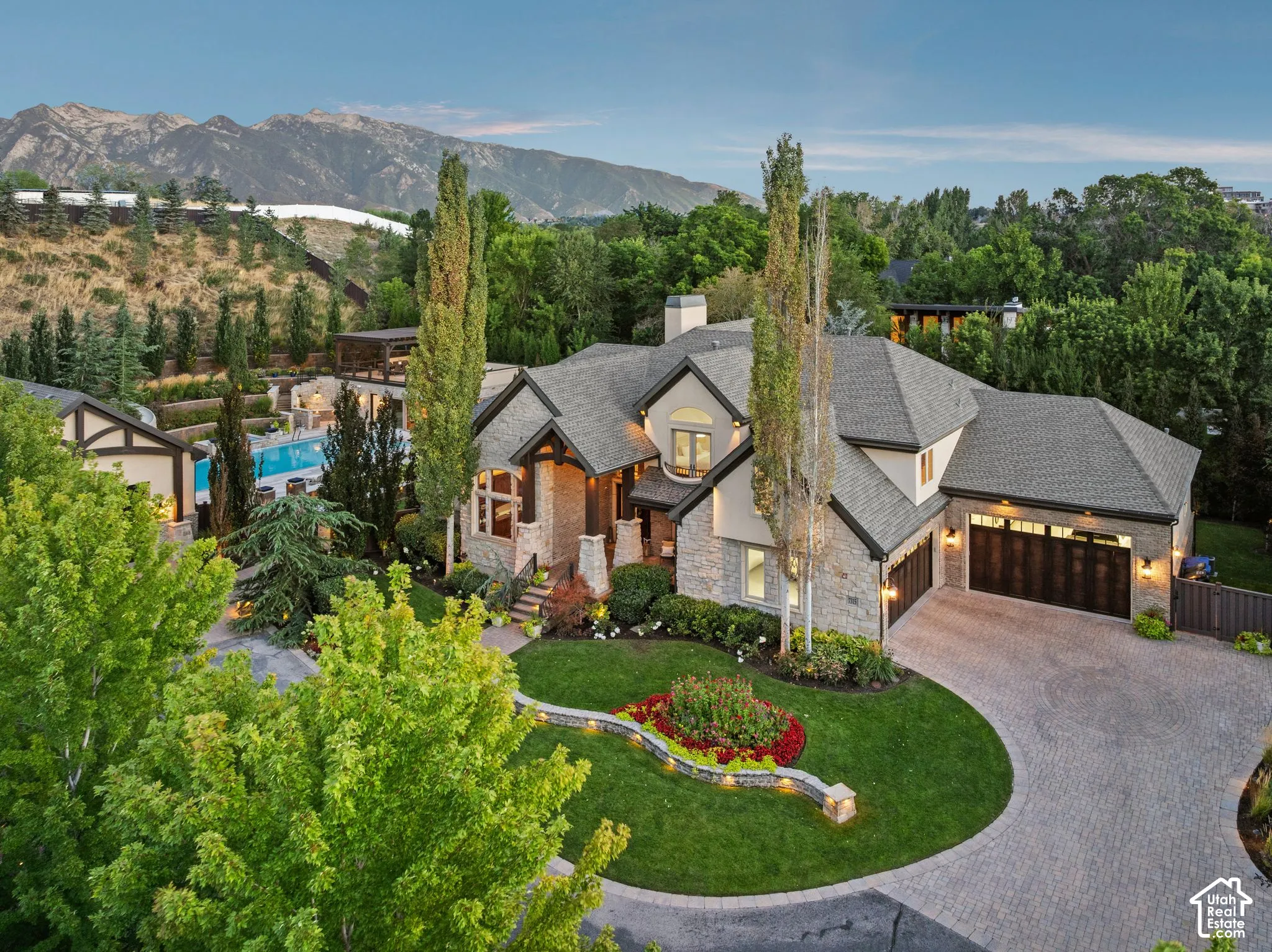 Stone and stucco exterior with multiple gables, three-car garage with wood-finish doors, circular paver driveway, manicured landscaping, and a terraced backyard with a swimming pool