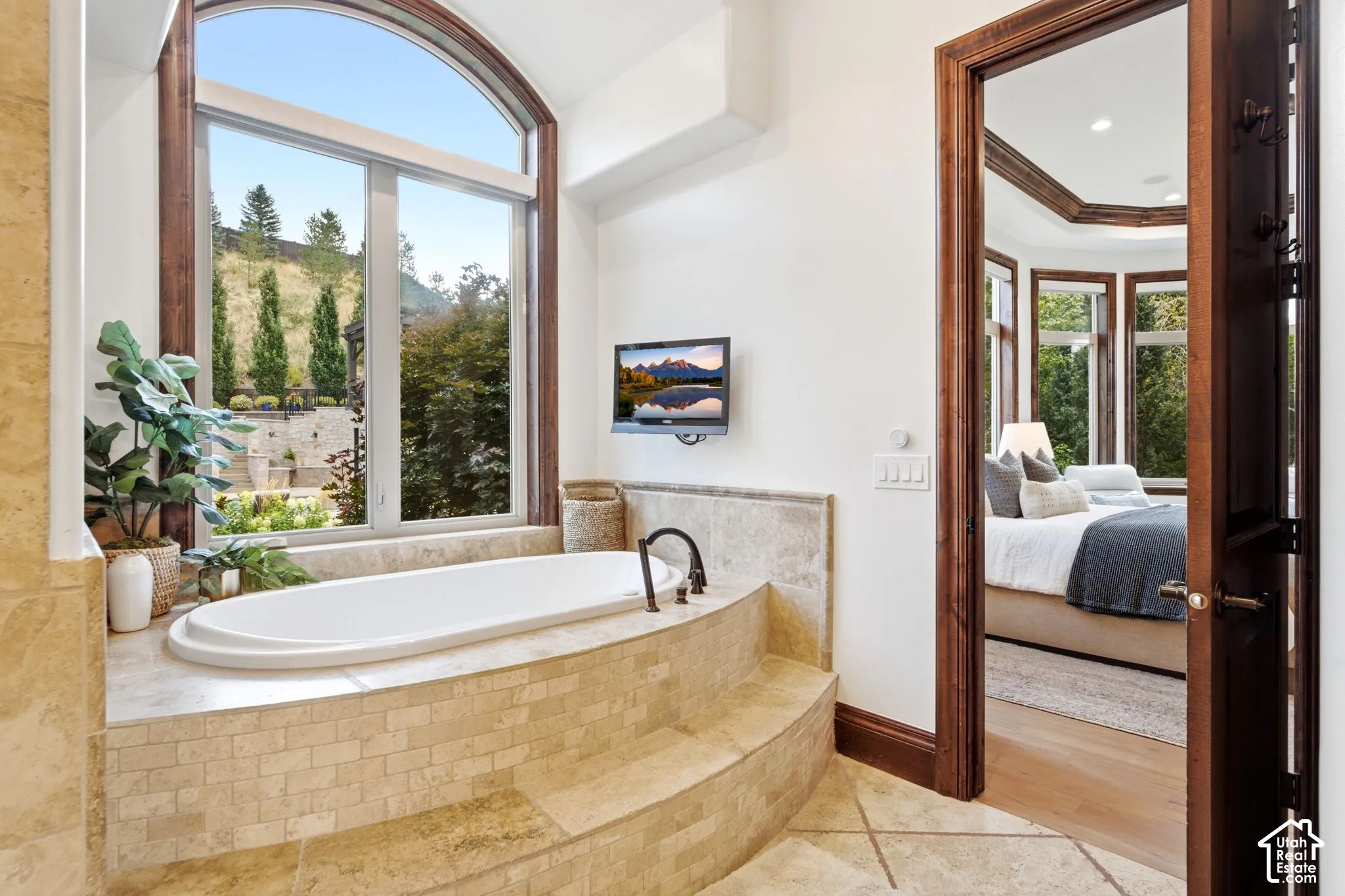 Soaking tub with tiled surround, arched window, wall-mounted television, and wood-framed doorway